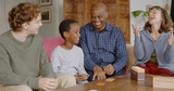 Two adults, a teenager, and a child play the Storymatic Classic storytelling card game around a table in a living room.