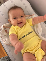 In a crib, a baby wears a yellow onesie that says "ex-superhero"